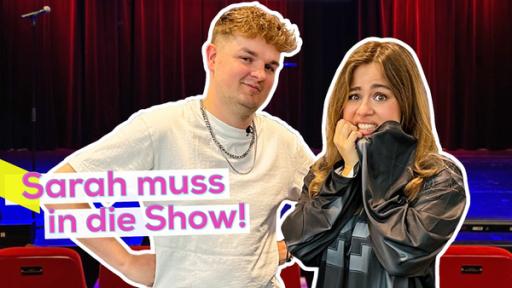 Rechte: KiKA/Jacqueline Kupey Comedian Tobii und Moderatorin Sarah stehen nebeneinander. Sarah mit ängstlichem Blick. Im Hintergrund die Bühne für den Auftritt. Aufschrift links: Sarah muss in die Show!