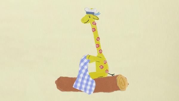 Eine Giraffe mit Mütze und Geschirrtuch auf einem Baumstamm. 