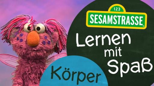 Der als Abby verzaubert Elmo mit Flügeln, Sommersprossen und rosanem Fell, steht neben einem Schild auf dem steht: "Sesamstraße: Lernen mit Spaß - Körper"