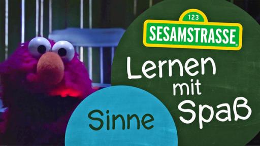 Elmo wird von unten angeleuchtet und steht neben einem Schild auf dem steht: "Sesamstraße: Lernen mit Spaß - Sinne".