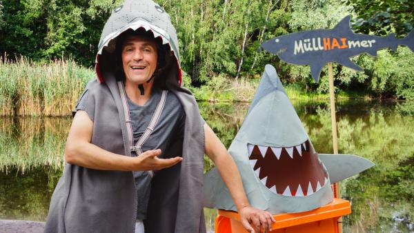 Fritz steht mit einem Haikostüm verkleidet am Seeufer, neben ihm eine Mülltonne als Hai | Rechte: ZDF