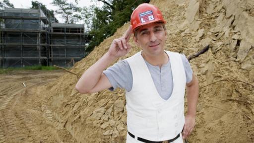 Fritz steht in Dachdeckerkleidung mit Bauhelm auf einer Baustelle.