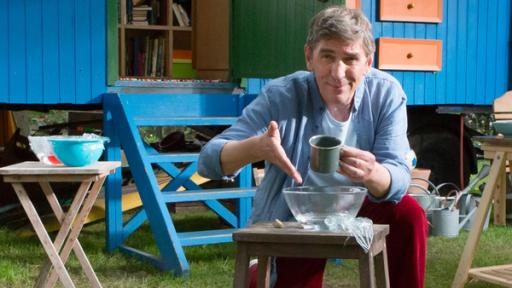 Fritz sitzt vor dem Bauwagen mit einer Tasse in der Hand und vor sich eine Schussel mit Wasser