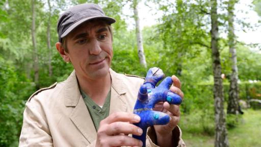 Fritz Fuchs im Wald hält einen blauen Gegenstand in der Hand.