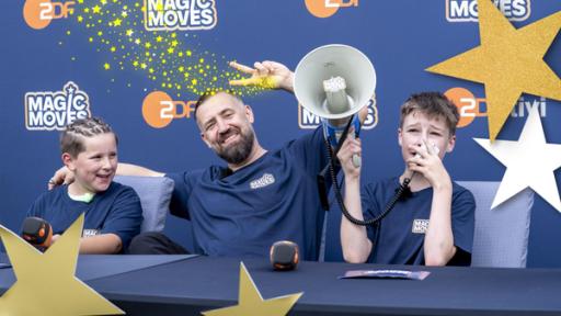 Gabriel (l.), Bürger Lars Dietrich (m.) und Maxi (r.) sitzen an einem Tisch. Gabriel (l.) hält ein ZDF-Mikrofon in der Hand, Maxim (r.) ein Megaphon. Im Hintergrund eine Wand auf dem das ZDF-Logo und das "Magic Moves"-