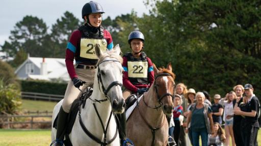 Natasha (links) und Caleb (rechts) reiten ihre Pferde beim Turnier gegen die Rivalinnen von Dulmoth Park. Beide tragen Helm, Schutzweste und ihre Startnummern. Im Hintergrund stehen einige Zuschauerinnen und Zuschauer.