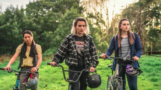 Caroline, Issie und Natasha stehen mit ihren Fahrrädern auf einer Wiese. An den Fahrradlenkern hängen ihre Helme.