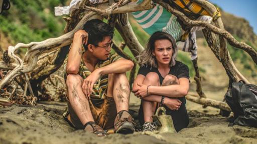 Issie (rechts) und Caleb (links) sitzen gemeinsam in einem selbst gebauten Zelt am Strand. Beide haben die Knie zum Oberkörper gezogen und sehen nachdenklich aus.