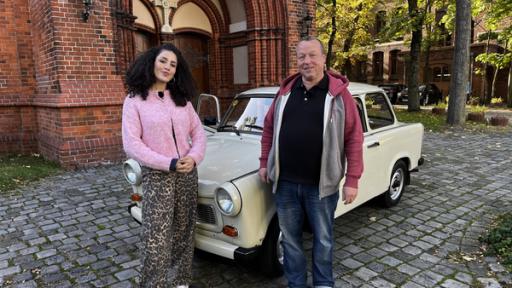 Reporterin Tessniem Kadiri und Zeitzeuge Michael Heinisch-Kirch stehen neben einem Trabi in Berlin vor der Erlöserkirche.