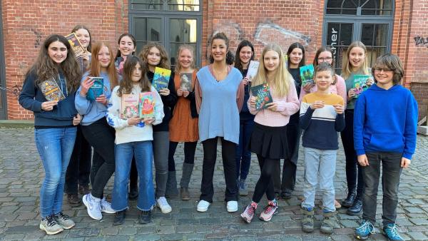 Auf einem Hof stehen mehrere Kinder, die Bücher ihren Händen halten und in die Kamera lächeln. In der Mitte der Gruppe steht die Moderatorin Tess. | Rechte: WDR/tvision