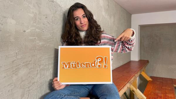 Mona sitzt auf einer Bank und zeigt auf ein Schild, auf dem "mütend" steht. | Rechte: WDR | tvision