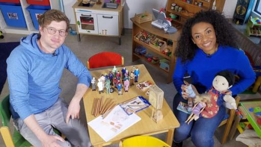 Robert sitzt mit Anne Chebu an einem Kindertisch, auf dem vielfältige und diverse Spielsachen liegen. Es gibt eine Schwarze und eine asiatische Puppe und Buntstifte in unterschiedlichen Hauttönen.