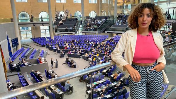 Reporterin Tessniem steht vor einer Glasabtrennung und schaut in die Kamera. Unter ihr ist der Plenarsaal mit Abgeordneten des Deutschen Bundestages. | Rechte: WDR | tvision