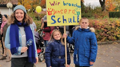 Gesa steht neben Leon (12) und Tanja (8). Die beiden Kinder halten ein Schild mit der Aufschrift "Wir brauchen unsere Schule".