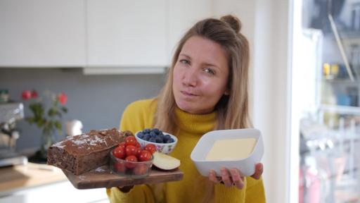 Jana schaut kritisch in die Kamera. Sie hält unterschiedliche Lebensmittel ins Bild, darunter Butter, Tomaten, Heidelbeeren, ein Brot und einen Apfel.