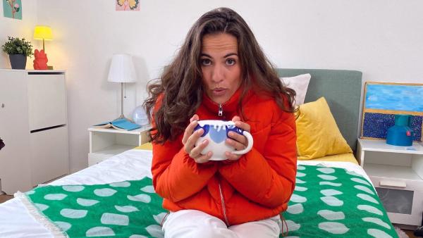 Mona sitzt in Winterjacke und mit einer großen Tasse Tee im Bett. | Rechte: WDR | tvision