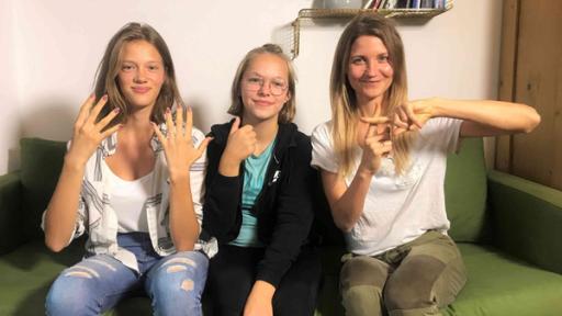 Jana sitzt mit der tauben Navina und ihrer Freundin Luana auf einer Couch. Sie zeigen in Gebärdensprache die Zahl neuneinhalb.