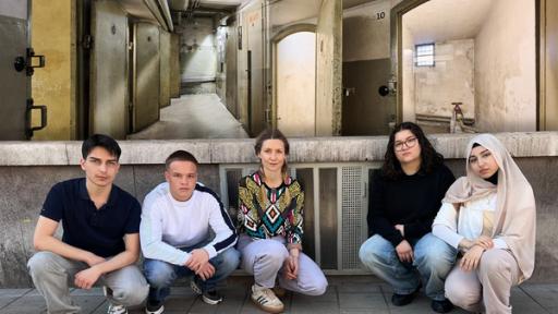 Reporterin Jana hockt zusammen mit vier Jugendlichen auf dem Boden vor einer Hauswand. Über ihnen ist ein Foto von Gefängniszellen projiziert, das aus dem NS-Dokumentationszentrum Kölns stammt.