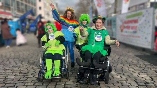 Reporterin Gesa steht neben Laki und Marie, die beide im Rollstuhl sitzen und deren Bruder Luca auf dem Kölner Rosenmontagszug.