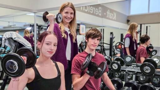 Reporterin Jana Forkel steht lächelnd in einem Fitnessstudio zwischen zwei Jugendlichen, Emilia und Dorian. Alle drei halten eine Kurzhantel in der rechten Hand.