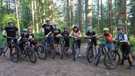 Gruppenfoto: Sieben Kinder, zwei Erwachsene und Reporterin Tessniem stehen mit Helmen und Mountainbikes im Wald und lächeln in die Kamera.