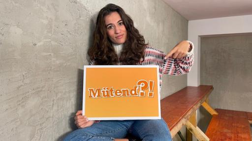 Mona sitzt auf einer Bank und zeigt auf ein Schild, auf dem "mütend" steht.