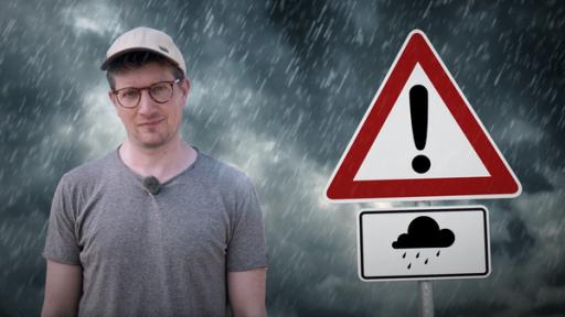 Reporter Robert steht vor einem Hintergrund mit dunkler Wolkenfront, aus der es regnet. Neben ihm ist ein Schild zu sehen, das vor Hochwasser warnt.