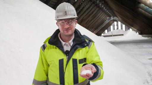 Robert steht in einer Halle, in der Salz aufgetürmt ist. Er trägt Schutzkleidung und hält Salz in der Hand.