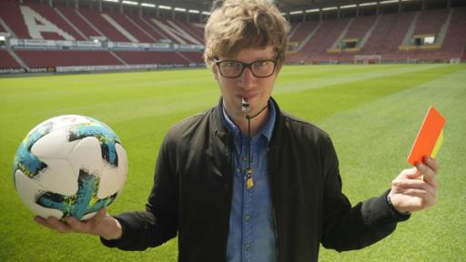 Robert auf dem Fußballfeld mit einem Fußball in der rechten und einer Roten/Gelben Karte in der linken Hand