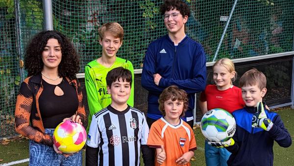 Tessniem hält einen Fußball in der Hand und steht neben einer Gruppe Kinder auf einem Fußballfeld. | Rechte: WDR | tvision