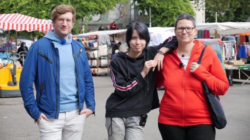 Reporter Robert steht mit dem 14-jährigen Alex und seiner Mutter Mel auf einem Wochenmarkt. Alex lehnt sich mit einem Arm auf die Schulter seiner Mutter.