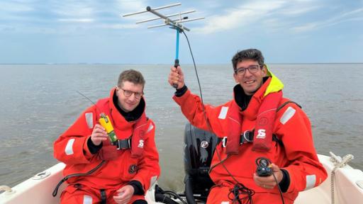 Reporter Robert und der Unterwasserlärm-Forscher Joseph "Jeff" Schnitzler sitzen in einem Boot. Robert hält einen Sender in der Hand und Jeff eine Antenne.