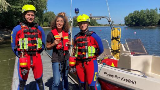 Reporterin Tessniem steht zwischen Kevin und Cécile auf einem Steg. Kevin und Cécile tragen Schutzkleidung.