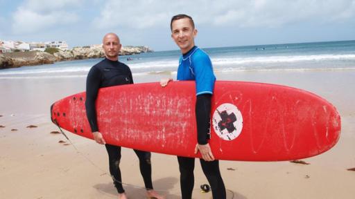 Eric steht zusammen mit Surflehrer Lars am Strand. Zusammen tragen sie ein Surfboard unter ihrem Arm. Im Hintergrund ist das Meer zu erkennen.