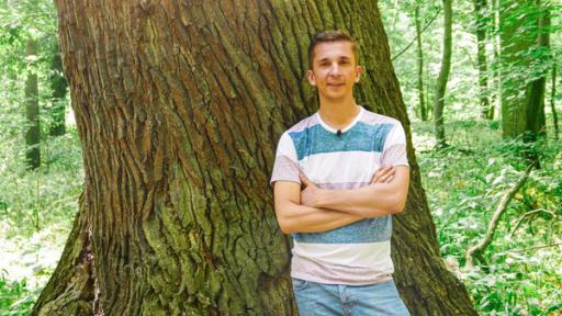 PUR+ Moderator Eric Mayer steht mit verschränkten Armen vor einem Baum. Zu sehen ist Eric vor einen sehr breiten Baumstamm.