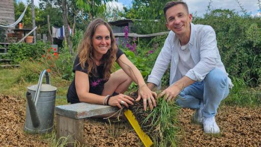 Moderator Eric Mayer kniet zusammen mit Permakultur Designerin Johanna Häger auf dem Boden eines kleinen Gartenfeldes. Beide halten einen Büschel Gras mit Wurzeln und Erde in der Hand. Beide blicken freundlich in die Kamera.