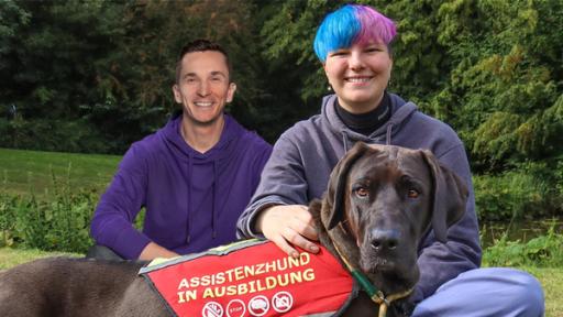 Assistenzhund Frieda liegt vorne im Bild in der Mitte und trägt ein Geschirr mit der Aufschrift "Assistenzhund in Ausbildung". Dahinter sitzt ihr Frauchen und links daneben Eric. Beide sitzen im Schneidersitz.