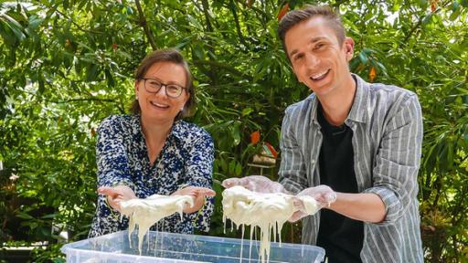 Eric Mayer (rechts) zusammen mit Wissenschaftsjournalistin Susanne Wedlich (links): Beide stehen an einem Tisch mit einem Becken voll selbst gemachtem Schleim aus Wasser und Maisstärke. Beide halten ihre Hände hoch die voll weißem Schleiß sind und schauen in die Kamera. 