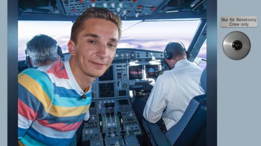 Über den Wolken: PUR+ Moderator Eric Mayer steht im Cockpit eines großen Flugzeugs. Er schaut durch die geöffnete Cockpittür in Richtung der Kamera. Im Cockpit sitzen Pilot und Copilot. Man sieht sie von hinten.