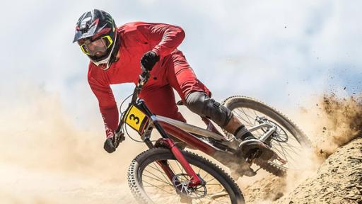 Ein Downhill-Fahrer im roten Anzug und mit Helm fährt auf einem Mountainbike einen Hügel hinab. Er scheint Teilnehmer eines Radrennens zu sein, da sein Fahrrad die Nummer Drei am Lenker trägt. Er wirbelt eine Menge Staub auf.