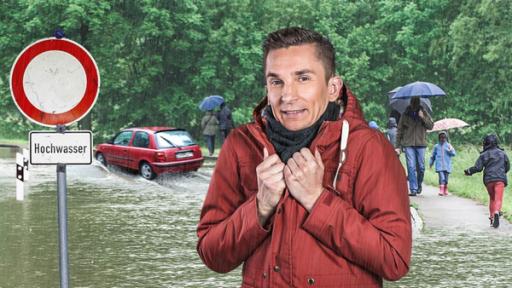 Moderator Eric Mayer steht mit angespannter Miene neben einem Schild mit der Aufschrift "Hochwaser". Im Hintergrund laufen Menschen mit Regenschirmen und es fährt ein Auto durchs Wasser auf einer überfluteten Straße.