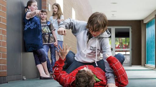 In einem Schulflur: Auf dem Boden liegt ein Junge. Ein anderer Junge kniet über ihm und scheint ihn schlagen zu wollen. Links an der Wand im Flur stehen zwei Schülerinnen und ein Schüler. Sie beobachten den Streit. Ein Mädchen zeigt mit der rechten Hand in Richtung der streintenden Jungen am Boden.