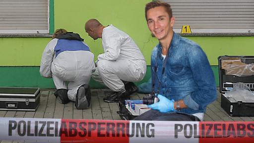 Eric Mayer kniet auf dem Gehweg vor einem Haus. Er trägt eine Fotokamera um den Hals und lächelt. Hinter ihm arbeiten kniend vor dem Haus zwei Personen in weißen Overalls und nehmen Spuren auf. Auf dem Gehweg liegen drei Koffer der Spurensicherung.