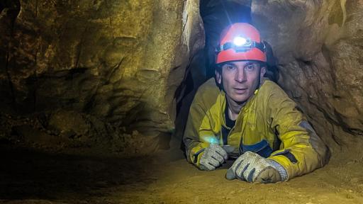 Moderator Eric Mayer liegt ausgestattet mit Handschuhen, einem gelben Overall und einem Helm mit Stirnlampe auf dem Boden einer Höhle in einer Höhlenspalte.