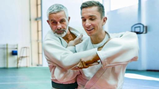 Moderator Eric Mayer im weißen Judo-Anzug steht vorne. Links hinter ihm steht einer der erfolgreichsten deutschen Judoka, Frank Wieneke, ebenfalls im weißen Judo-Anzug. Eric hält Trainer Frank am Arm und am Anzug, als würde er ihn über die Schulter zum Stürzen bringen wollen.