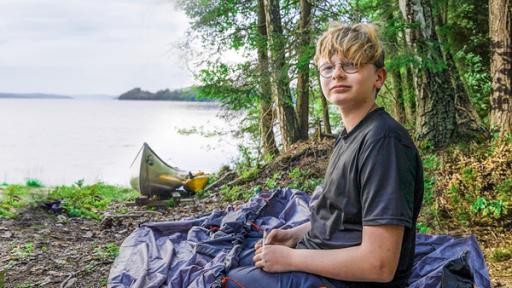 Marc sitzt auf einem steinigen Untergrund am Rande eines großen Sees. Neben ihm liegt das noch nicht aufgebaute Zelt. Ein paar Meter weiter liegt ein Kanadier am Ufer. Hinter dem See liegen bewaldete Berge.