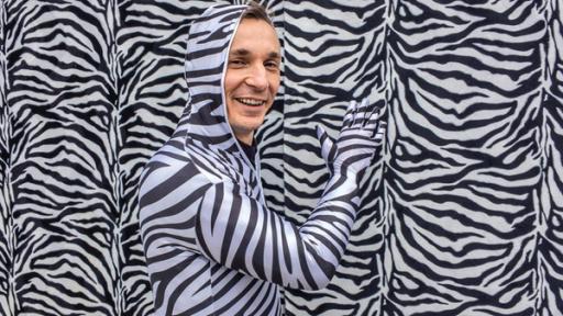 PUR+ Sendung Tarnung: Moderator Eric Mayer steht in einem Ganzkörper-Kostüm im Zebralook vor einer Wand im Zebralook. Nur sein Gesicht trägt keine Zebrastreifen und ist ungeschminkt.