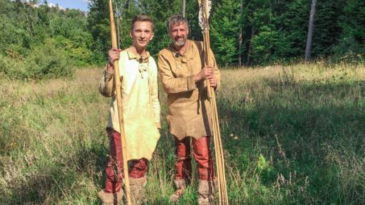 Moderator Eric Mayer steht links im Bild. Er trägt steinzeitiche Kleidung aus Leder und Lederstiefel. In der rechten Hand hält er einen riesigen Speer aus Holz. Rechts nebem ihm steht ebenso gekleidet und mit riesigen Pfeilen in der Hand ein Steinzeitexperte mit ihm auf einer Wiese.