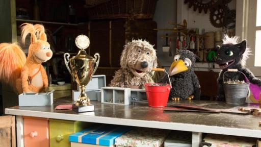 udis Rasselbande ist in der Garage um Rudi herum versammelt und schaut ihn angespannt an. Rudi greift mit dem Schnabel den Henkel eines vollen Eimers. Vor ihnen steht ein großer Pokal auf dem Tisch. 