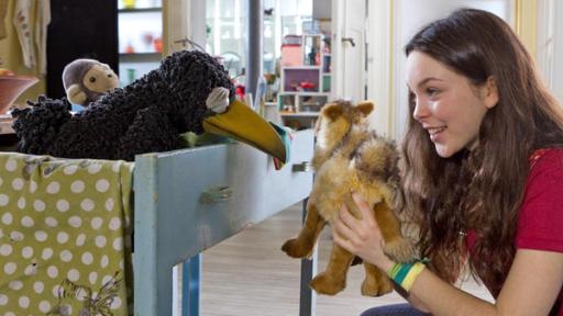 Das Mädchen Svea zeigt dem Raben Rudi, der auf dem Küchentisch hockt, ein Stoffkamel.